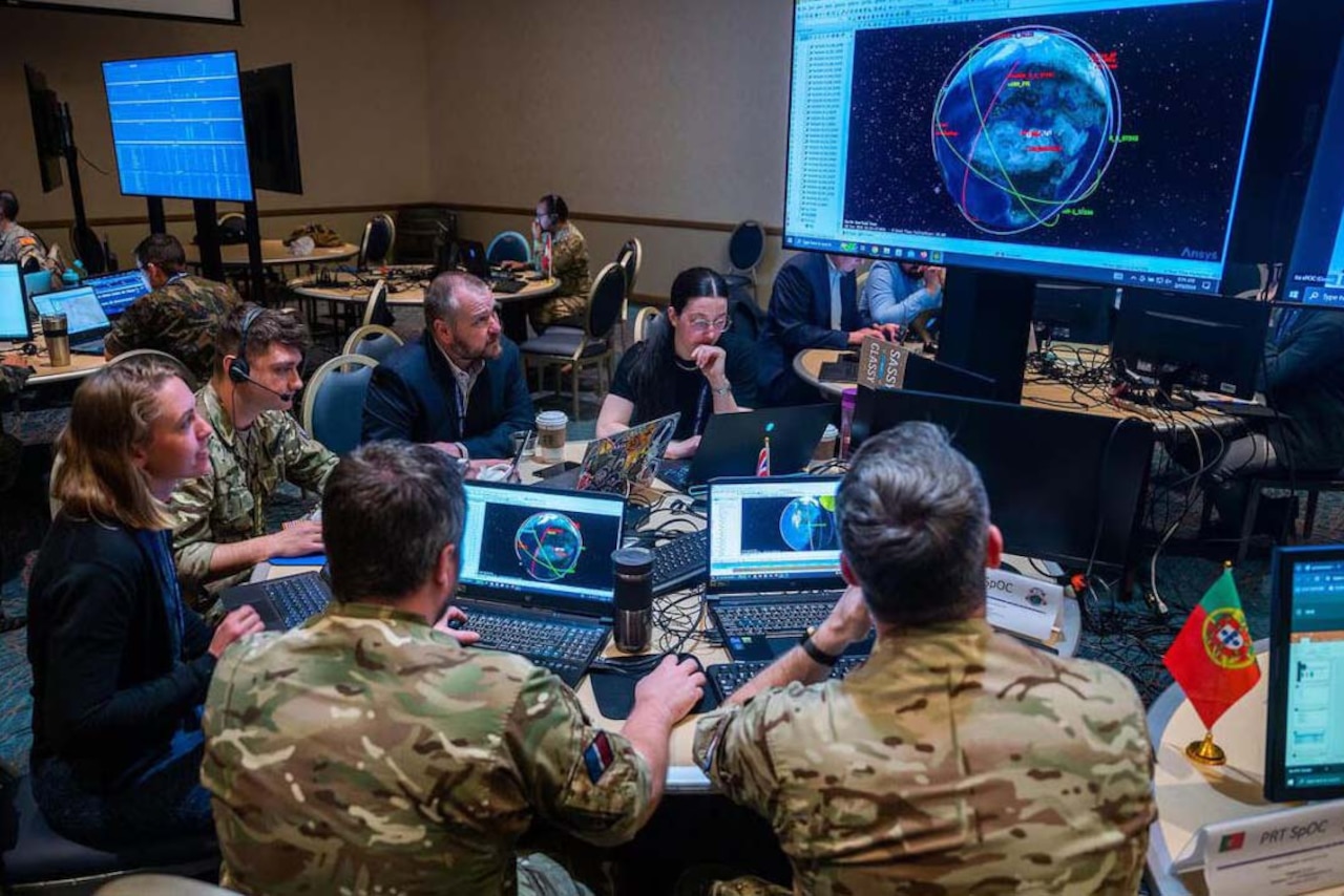 A group of civilians and foreign military personnel look at a computer terminal while seated.