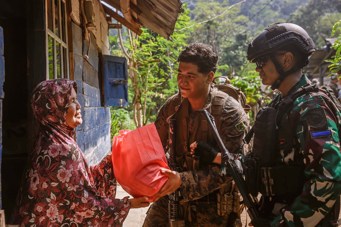 A U.S. and an Indonesian marine deliver a bag to a person at their door with trees in the background.