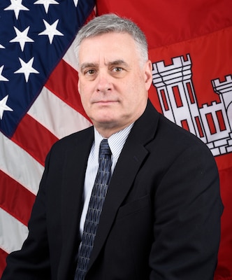 A man looks toward the camera. Behind him are the American and U.S. Army Corps of Engineers flags.