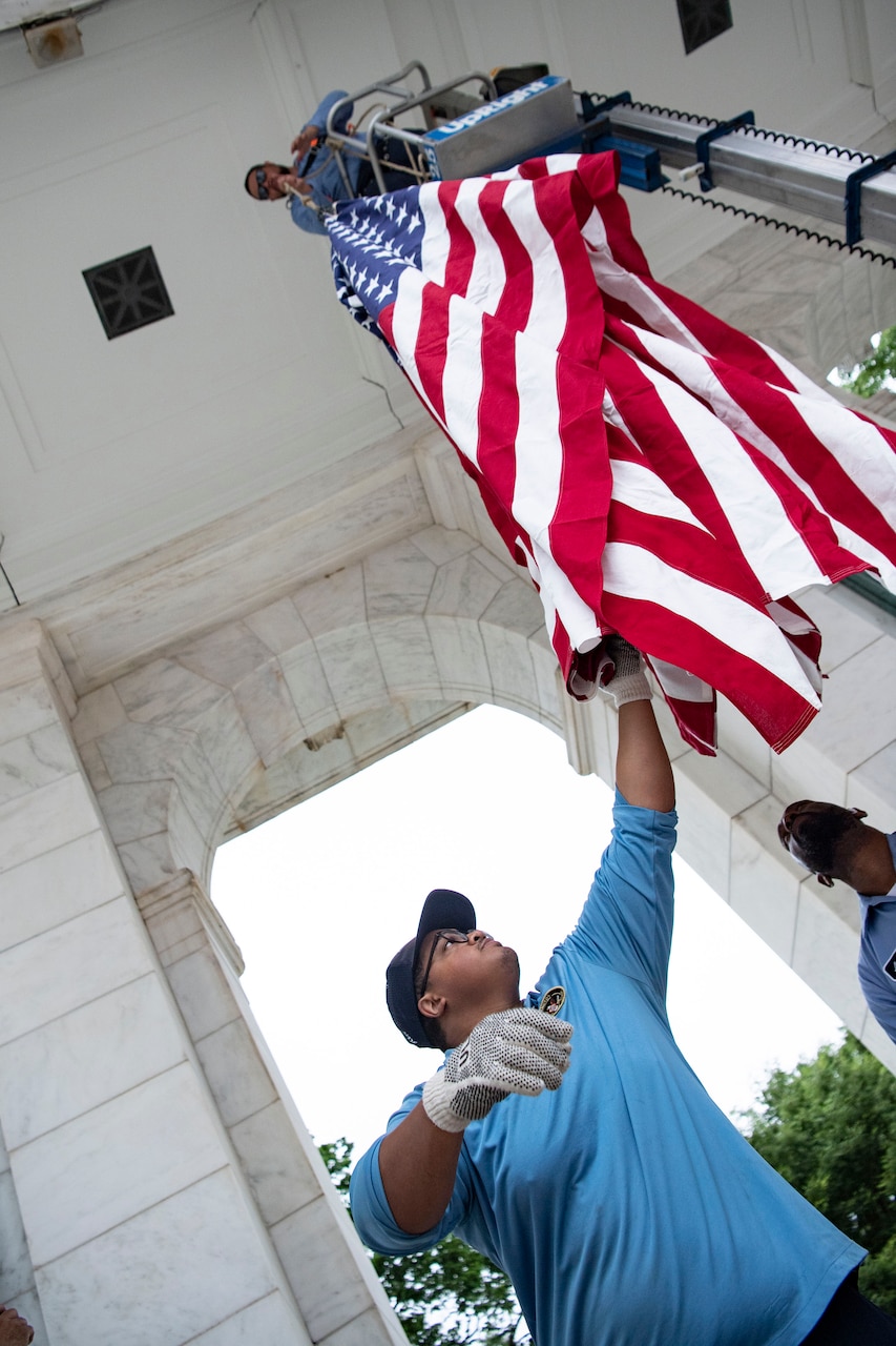 View from blow of a civilian in a raised platform holding one end of a U.S. flag at the top of a white building as another holds the other end below.