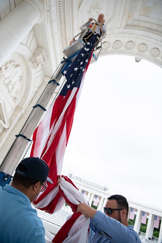 A civilian in a raised platform holds one end of a U.S. flag at the top of a white building as two others hold the other end below.