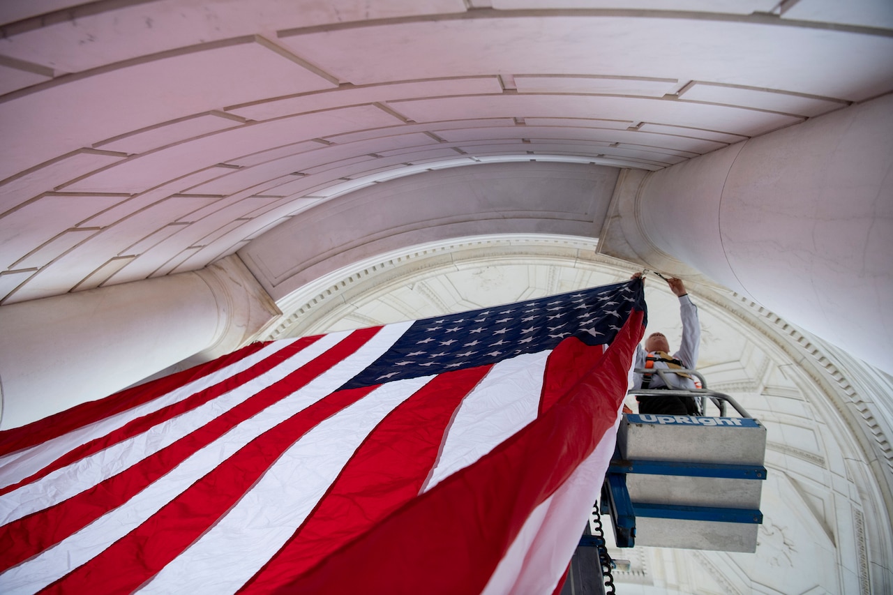 View from below of a civilian in a raised platform hanging a U.S. flag between white columns of a curved structure.