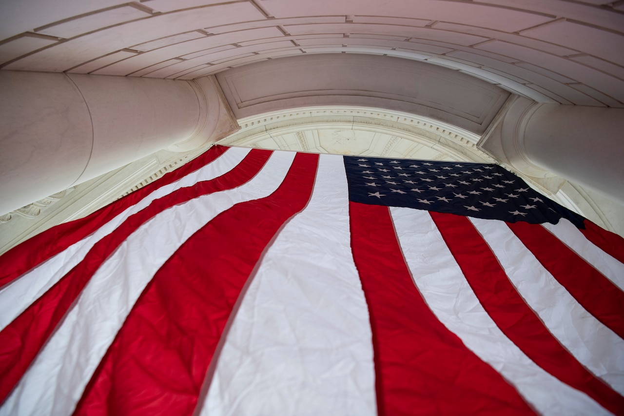 Angled view from below of a U.S. flag hanging between white columns of a curved structure.
