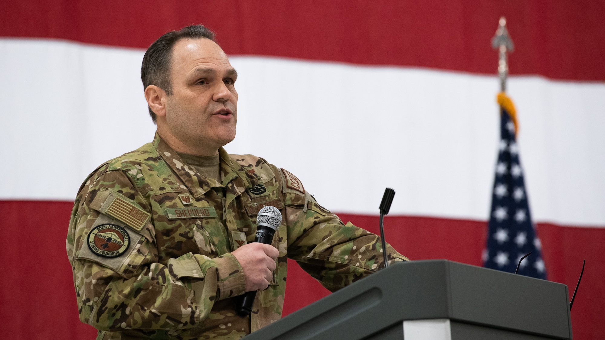 A man in U.S. Air Force uniform stands at a podium holding a microphone with an American flag banner in the background.