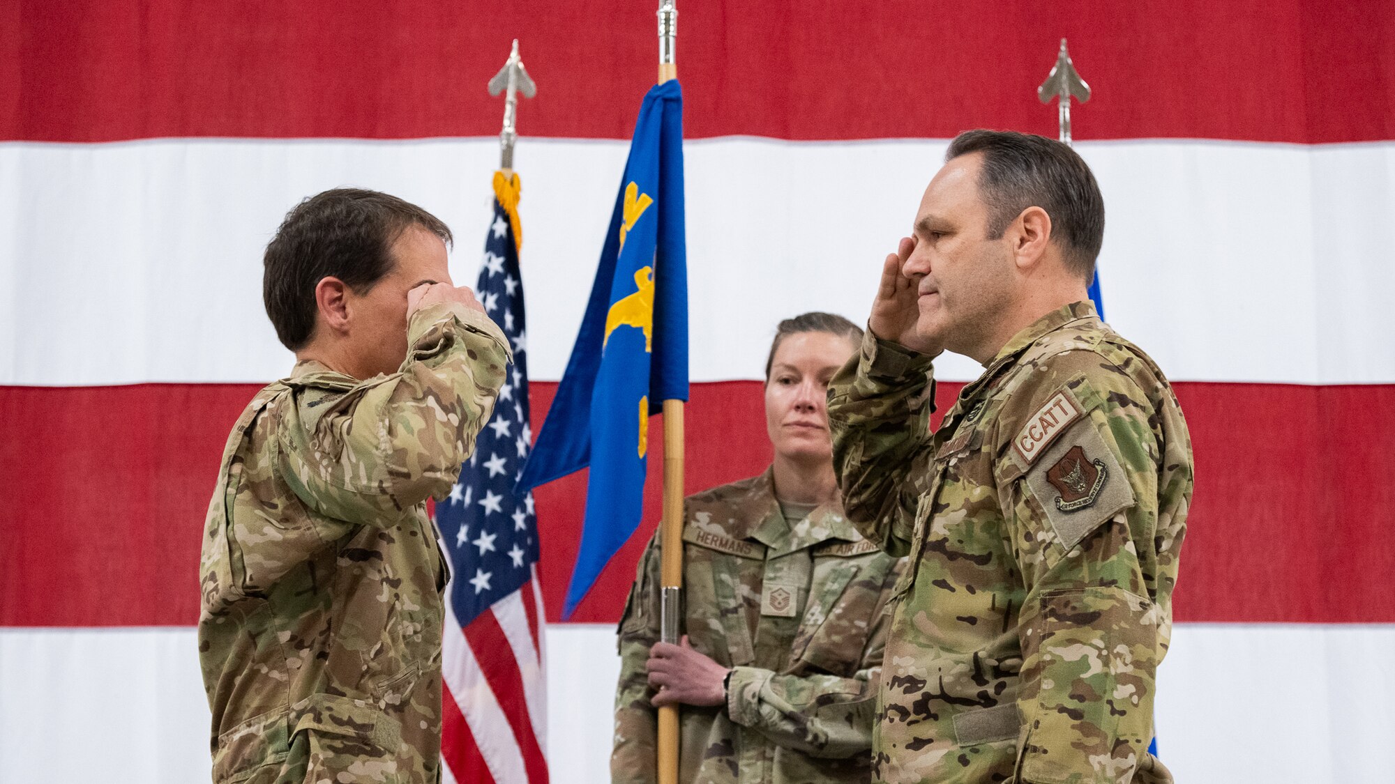 Two men in U.S. Air Force uniform salute one another while a woman wearing a U.S. Air Force uniform holds a flag between them.