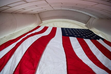 Angled view from below of a U.S. flag hanging between white columns of a curved structure.