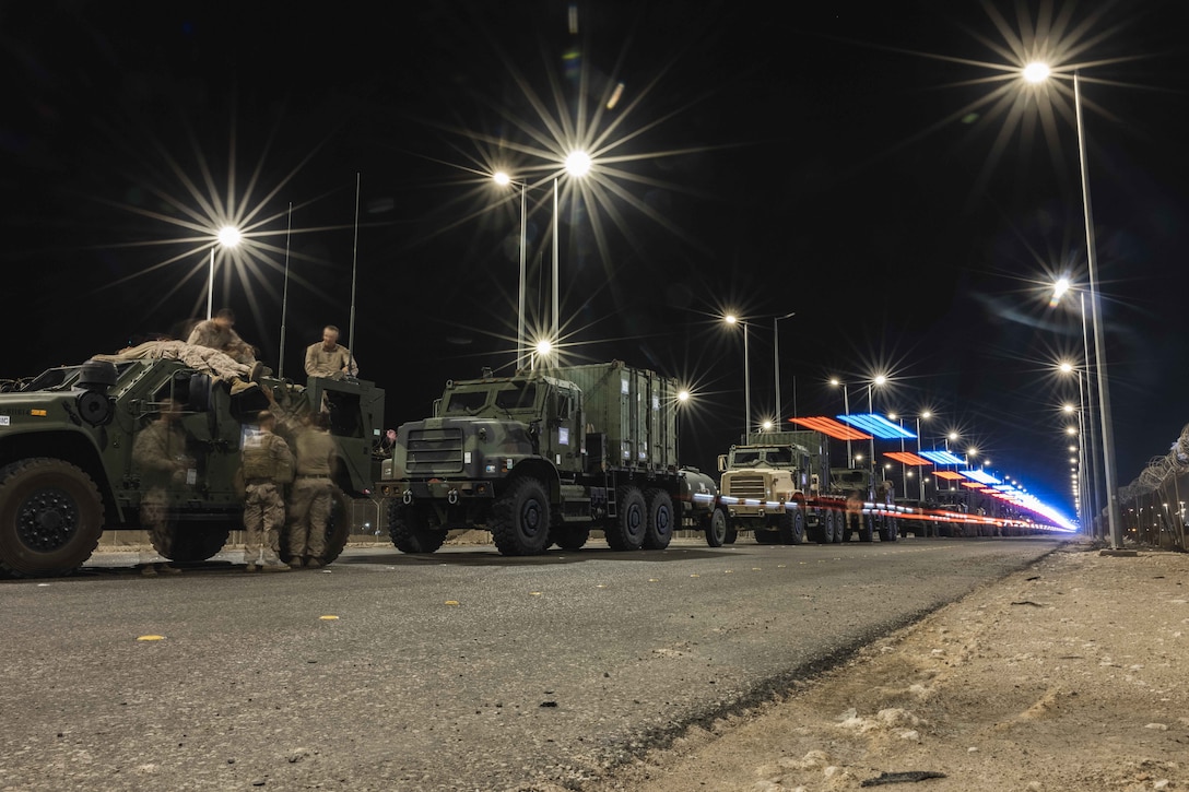 Military vehicles line up on a road lit up by streetlights and overhead lights in a desert setting at night.