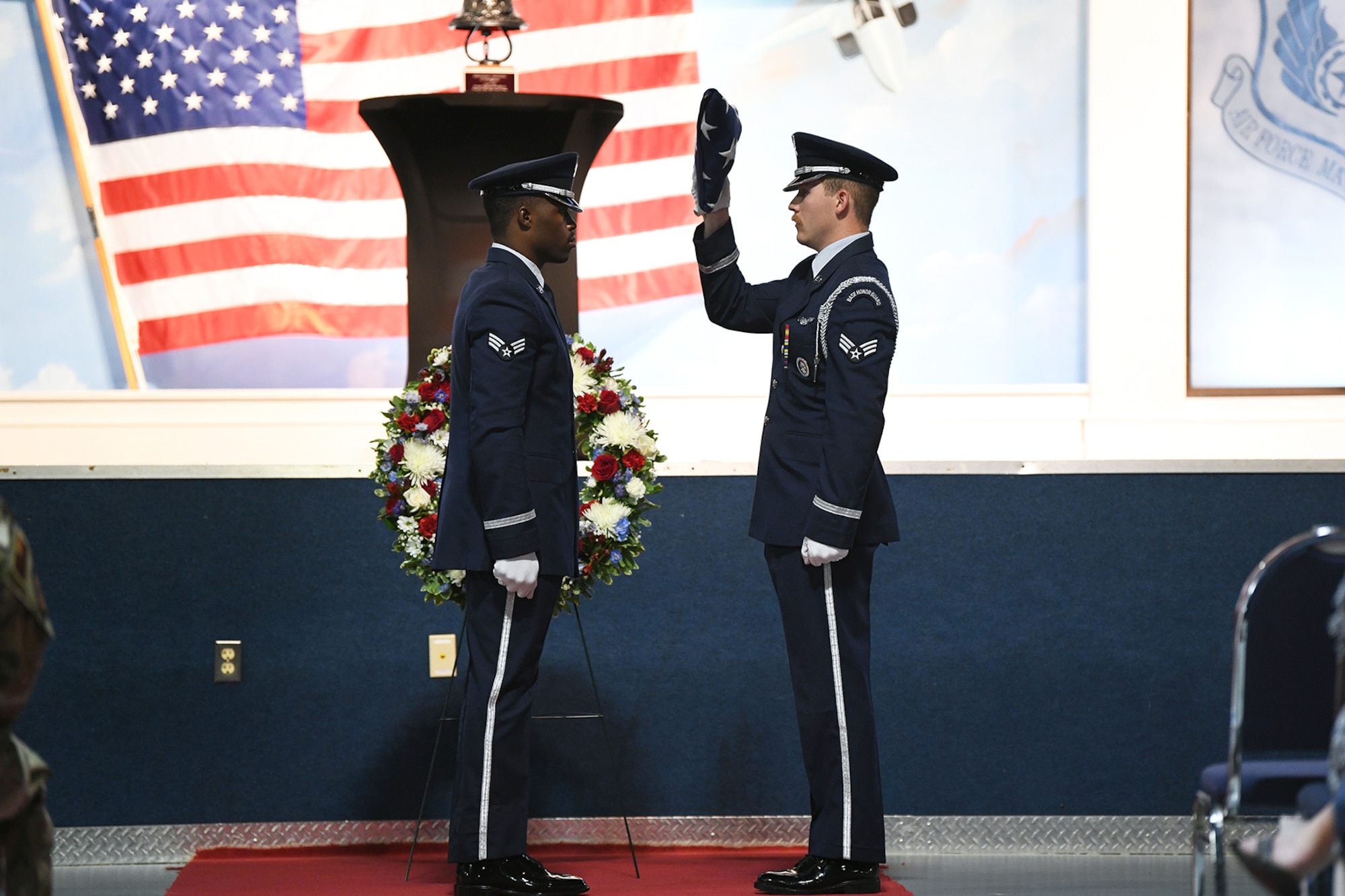 Two men in military uniform stand facing each other while man on the right lowers a U.S. flag folded into a triangle shape