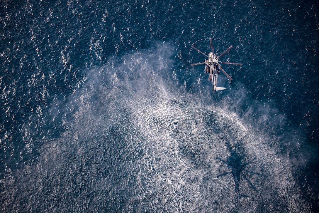 Aerial view of a military helicopter flying over open water and casting a shadow in its wake.