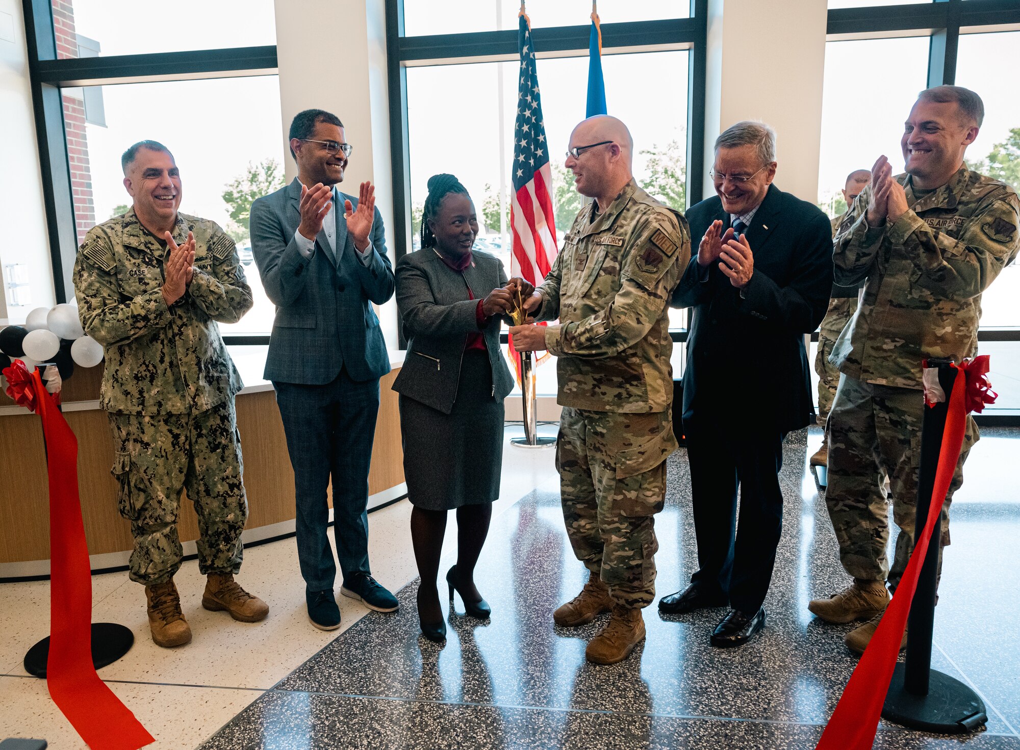 A group of men and women, civilians and military members, clap while the two in the middle hold a large pair of scissors