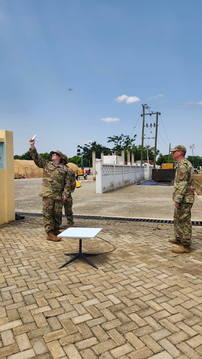 Two uniformed U.S. Air Force Airmen stand near a piece of a equipment on a sidewalk while a third raises a phone into the air.