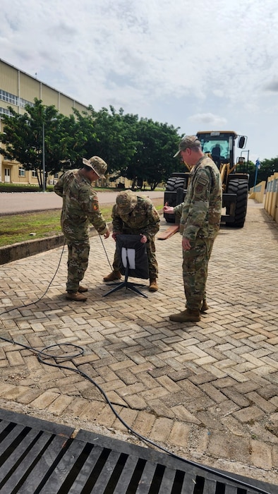 Three uniformed U.S. Air Force Airmen configure a piece of equipment on a sidewalk.