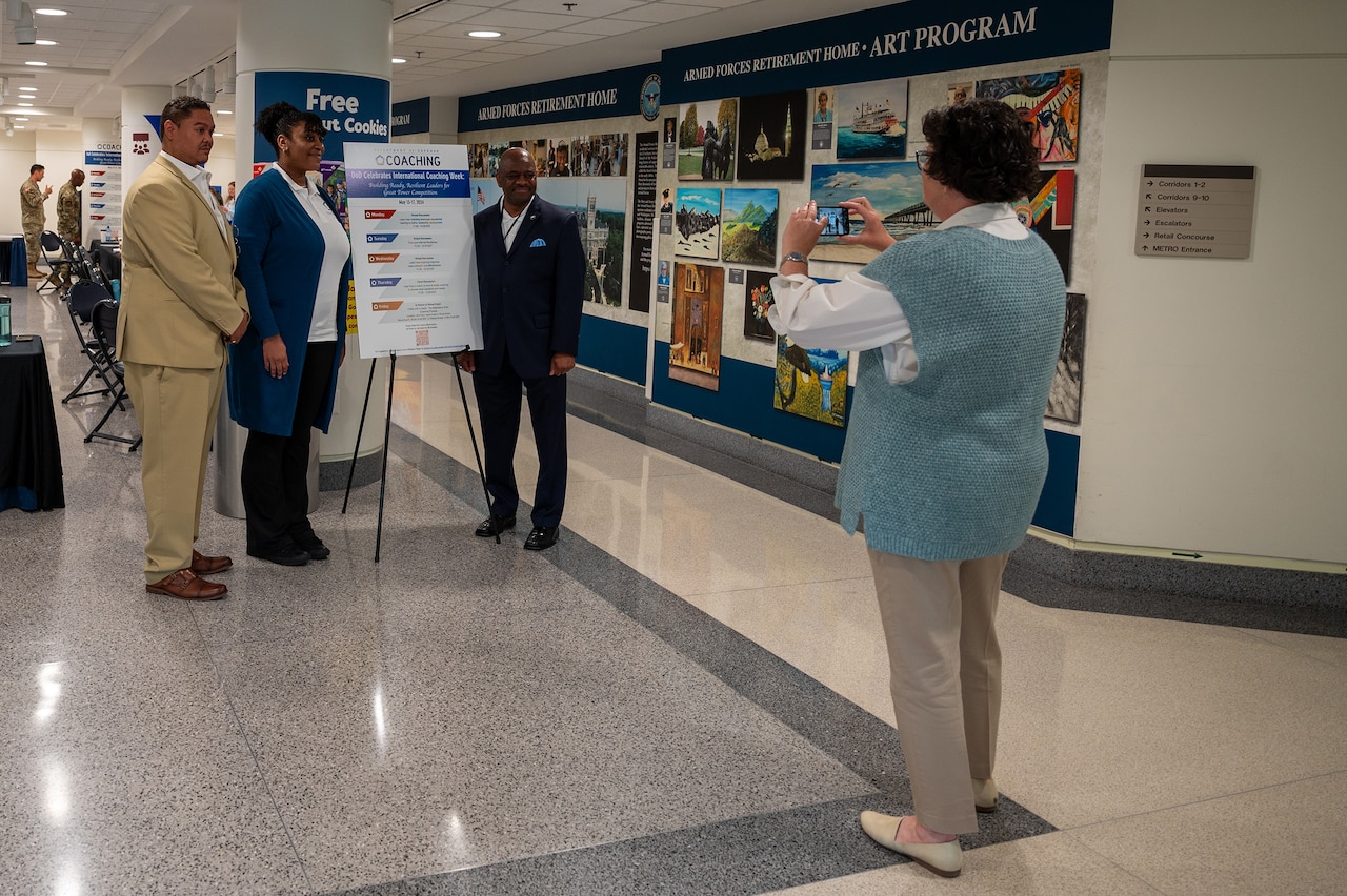 Three people wearing civilian attire pose for a photograph next to a sign celebrating International Coaching Week.
