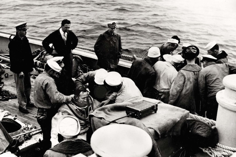 Two groups of men on a ship work to put diving suits on two other men.