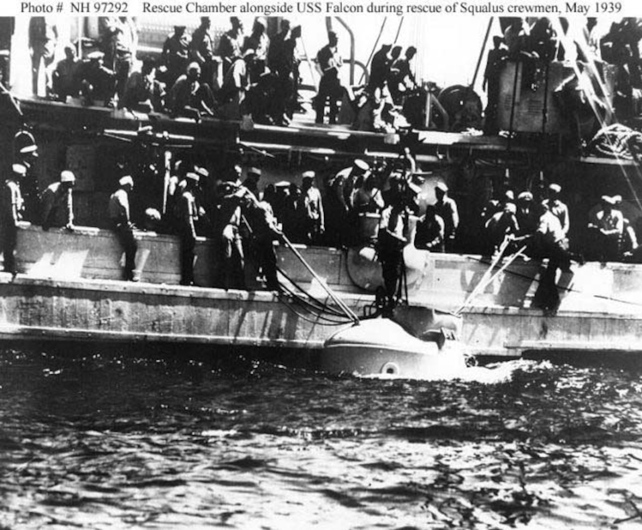 Several men on a ship use ropes to pull a large chamber out of the ocean.