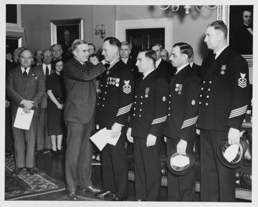 A man places a medal around another man’s neck. Three other men stand in line beside them. Others watch from a distance.