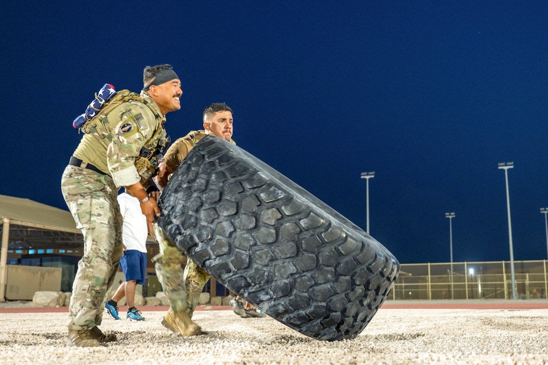 Two airmen lift a tire on a rocky surface near a tennis court.