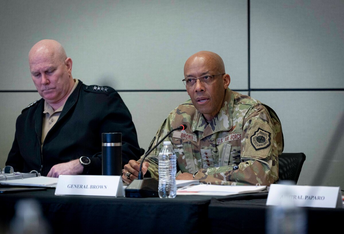 A man in military uniform speaks into a microphone. Another man sits beside him.