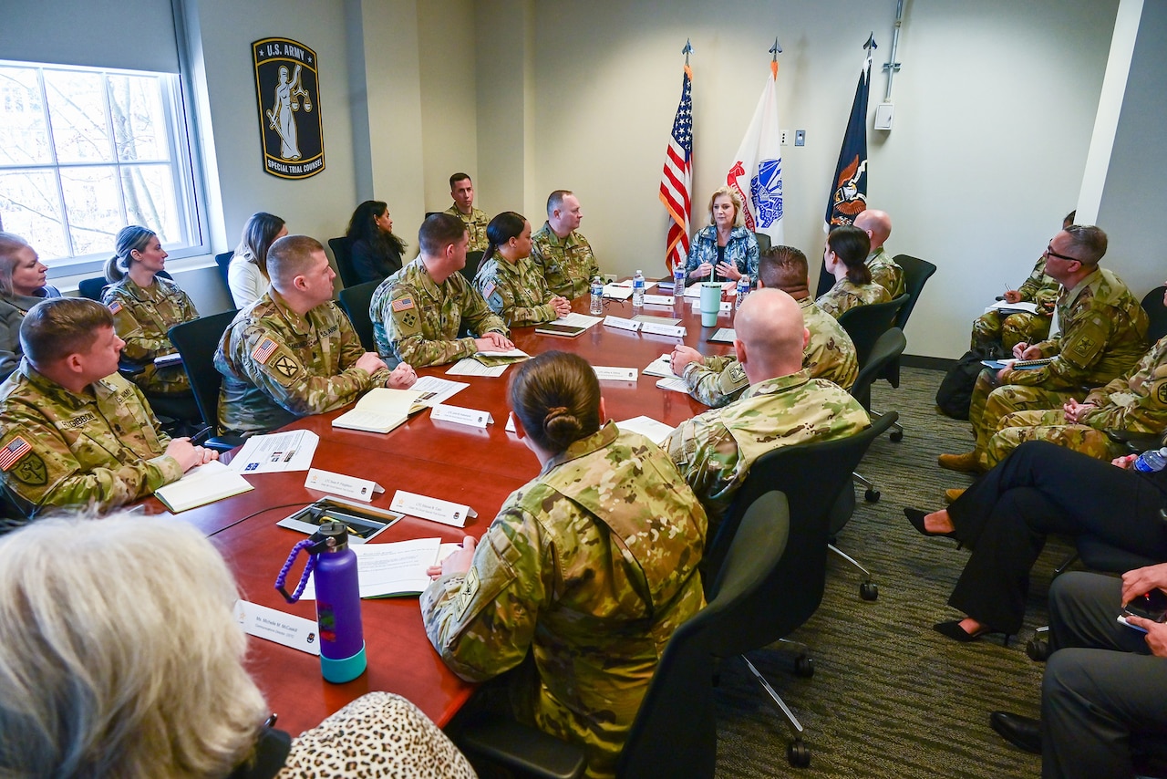 Service members in uniform are seated at a long table. A civilian is seated at the head of the table.