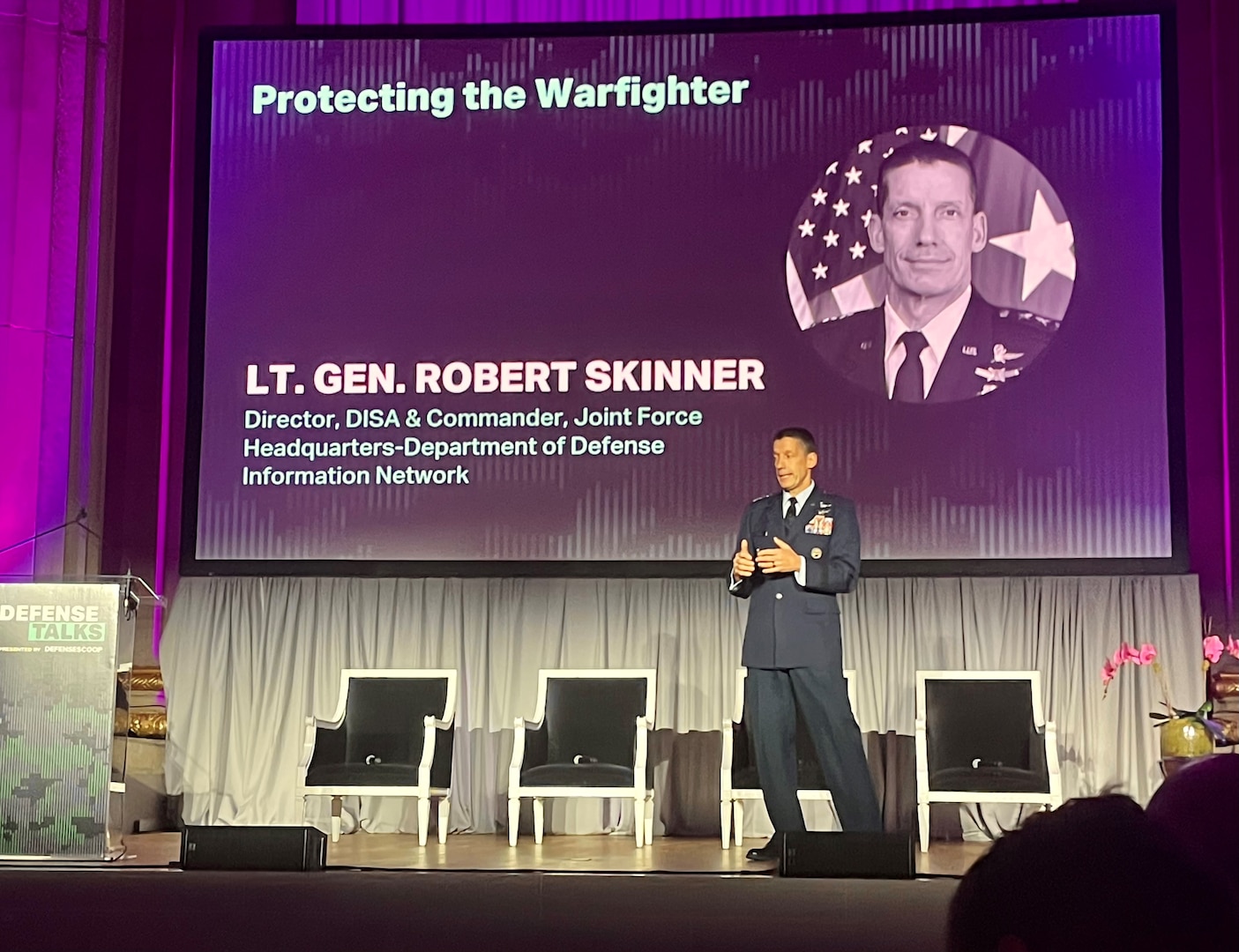 A man in a dress military uniform stands on a stage talking and gesturing with both his hands.