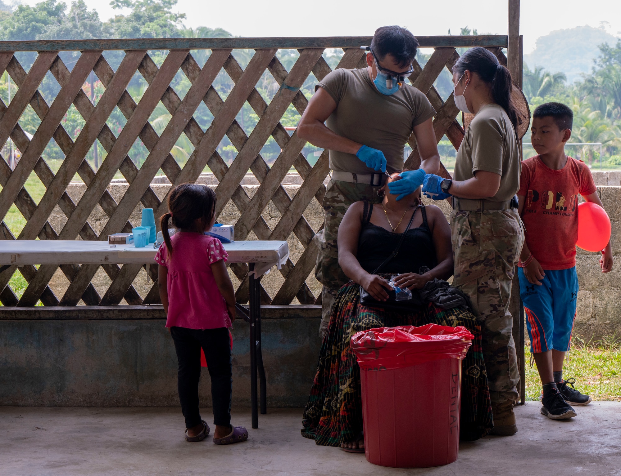 A medium photo of a military medical professional and his technician extracting teeth while two children watch.
