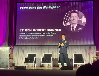 A man in a dress military uniform stands on a stage talking and gesturing with both his hands.