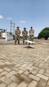 Three uniformed U.S. Air Force Airmen stand on a sidewalk behind a white flat object mounted on a stand.