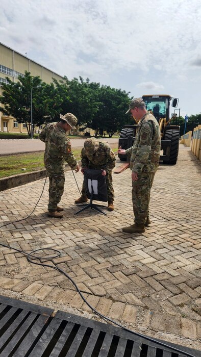 Three uniformed U.S. Air Force Airmen configure a piece of equipment on a sidewalk.