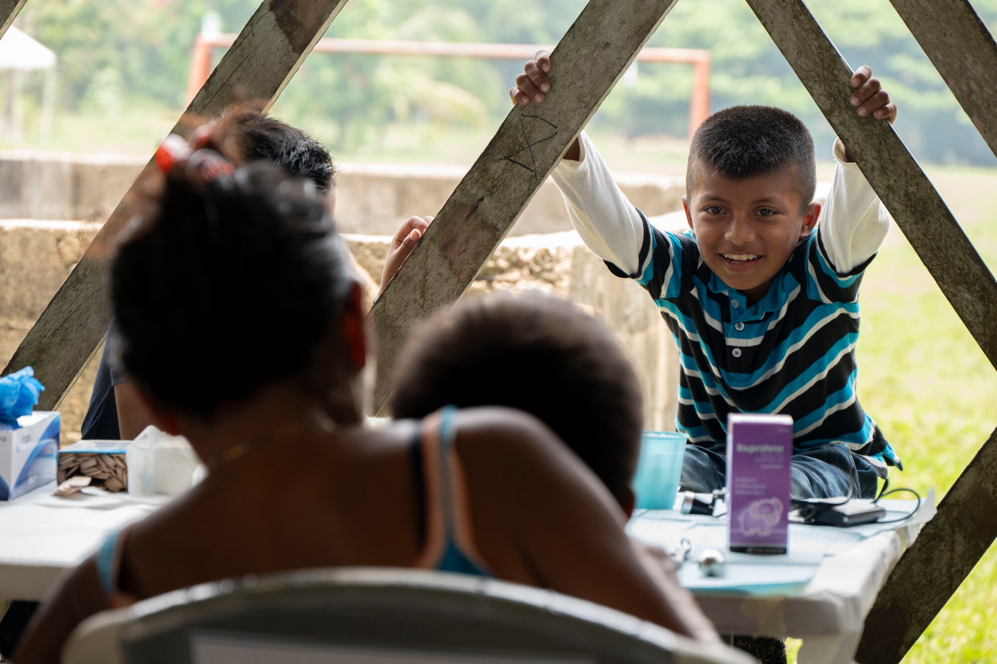 A photo of a mom with a boy sitting on her lap facing away from the camera while a little boy hangs on the building smiling at the mom facing the camera.