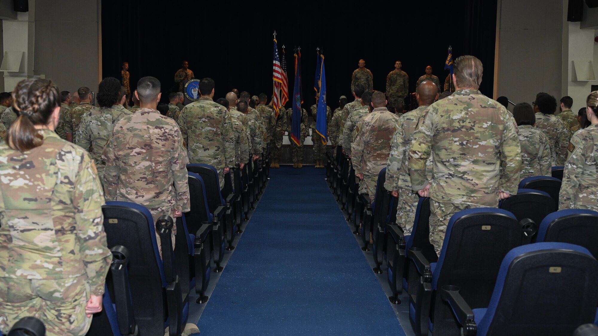 The 36th Maintenance Squadron Airmen stand at attention in front of their seats facing the stage.