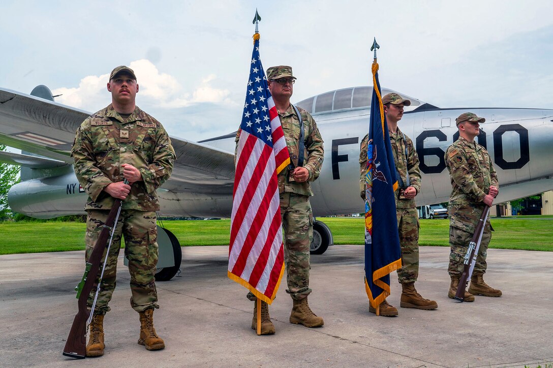 Four Air National Guardsmen stand in row, two holding flags, on a paved lot in front of a historic aircraft.