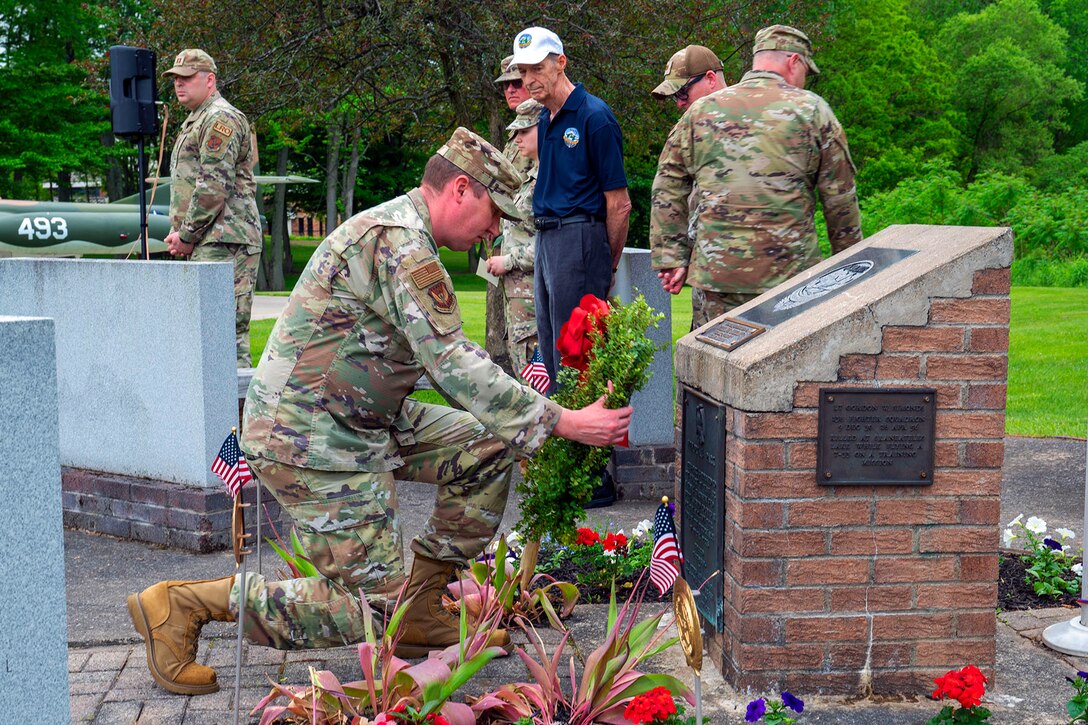 An airman kneels in front of a brick memorial to place a wreath as others stand nearby.