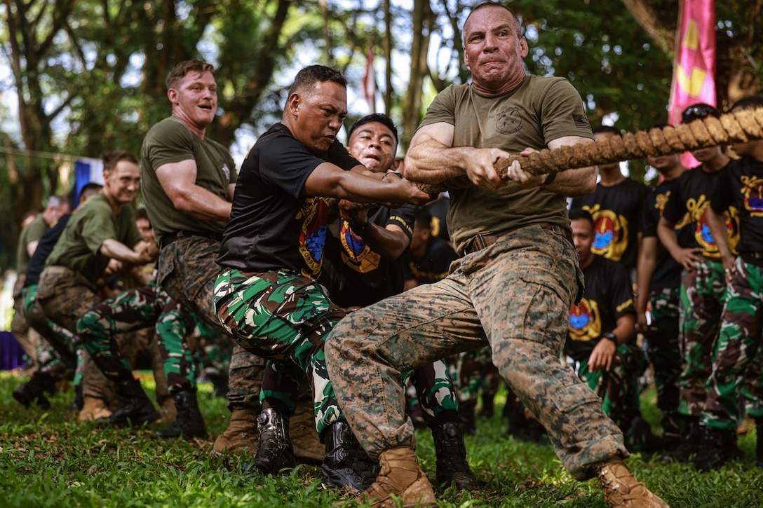 Marines grimace and smile as they pull on a thick rope in a jungle setting. Other service members watch