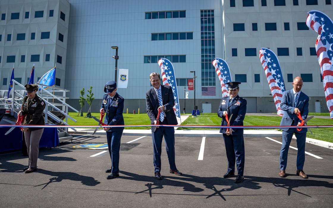 Five people, military and civilian, holding giant scissors prepare to cut a red, white, and blue ribbon