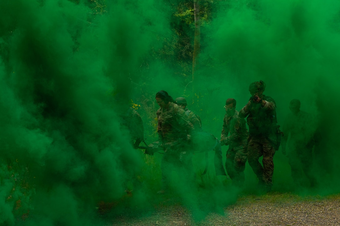 A group of soldiers carry a rescue stretcher through a wooded area surrounded by green smoke.