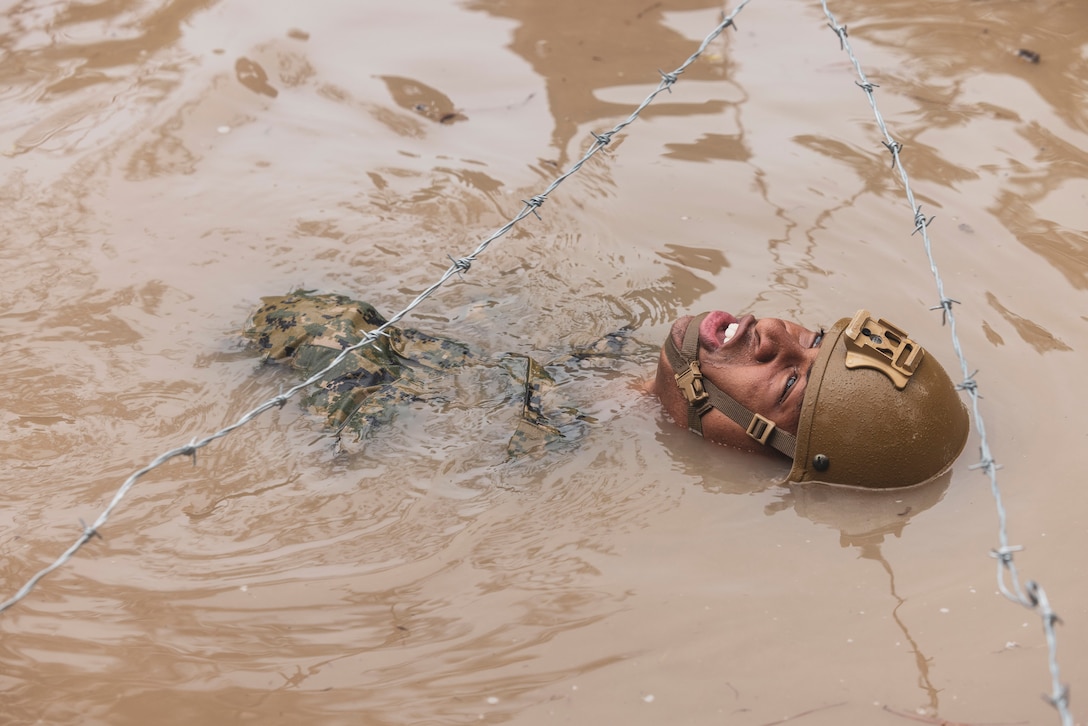 A U.S. Naval Academy midshipman wearing a helmet floats in muddy water underneath barbed wire.