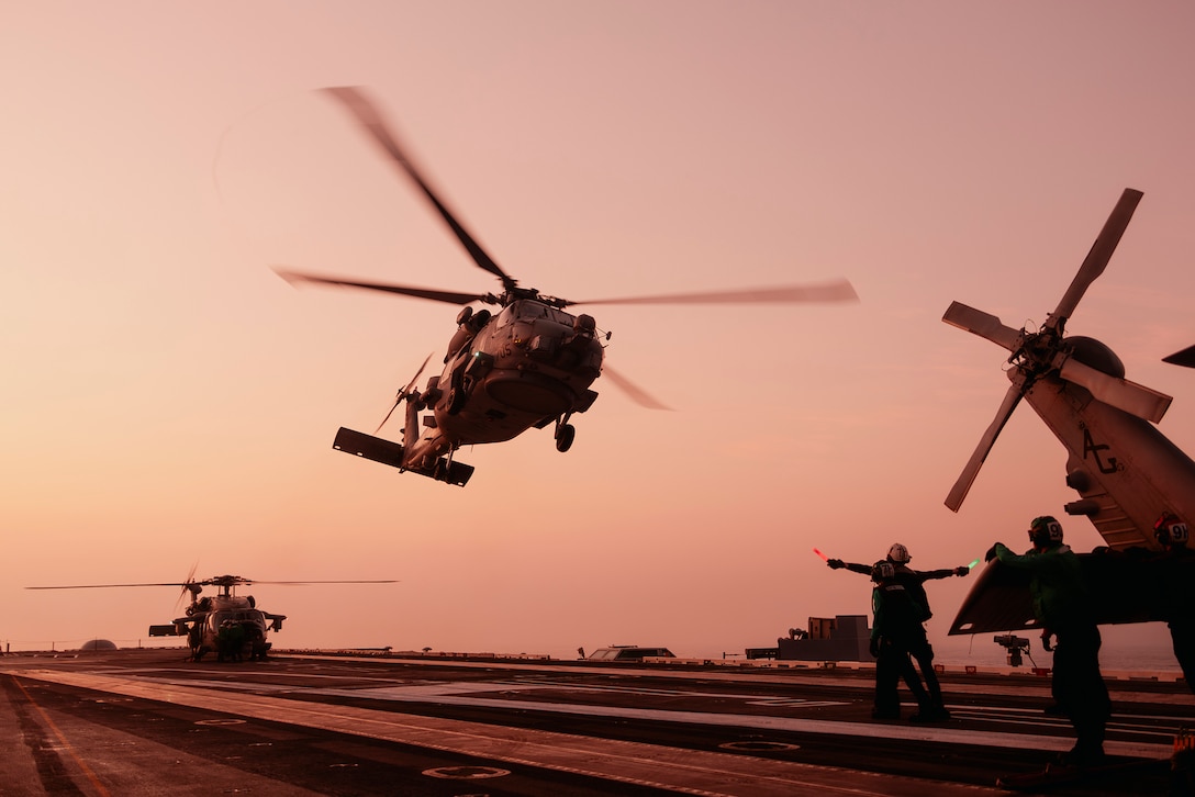A sailor signals as a Navy helicopter takes off from the flight deck of a ship against a dusty orange sky.