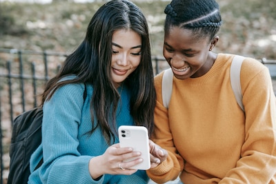 Two young women sit on a bench smiling and looking at a smartphone.