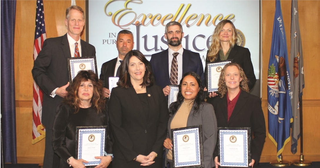 A group of professionally dressed people holding awards in two rows in front of a text screen surrounded by flags in the background.