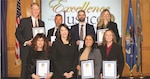 A group of professionally dressed people holding awards in two rows in front of a text screen surrounded by flags in the background.