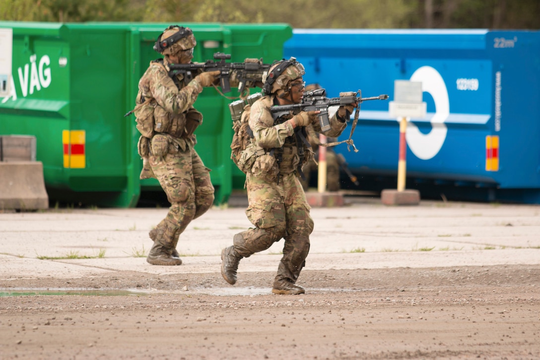 Soldiers aim weapons while moving across pavement with dumpsters in the background.