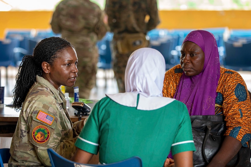 A soldier speaks with two people while they sit at a table as two fellow soldiers gather near rows of empty seats in the background.