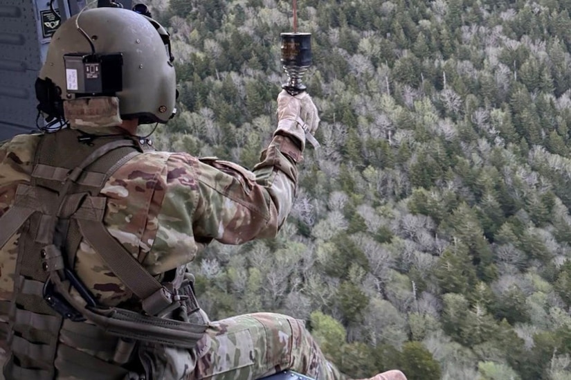 A soldier sits on the edge of a helicopter looking out over a forest.