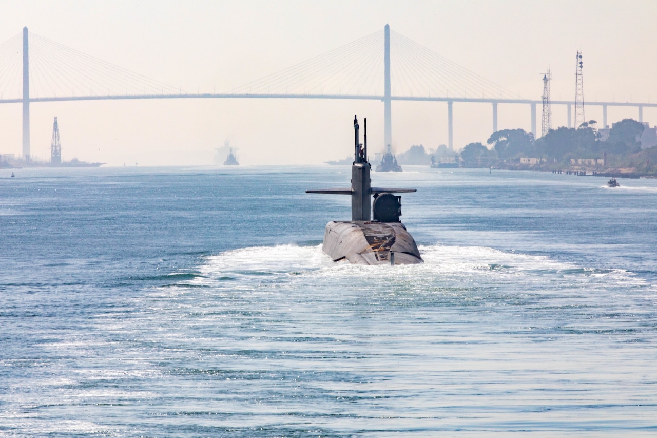 A submarine cuts through the water. In the background is a bridge.
