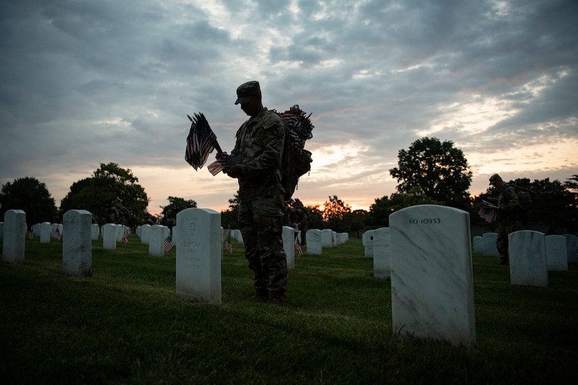 A soldier places U.S. flags at Arlington National Cemetery gravesites at twilight.