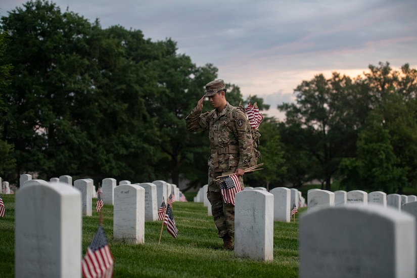 A soldier salutes a gravesite at Arlington National Cemetery while holding small U.S. flags.