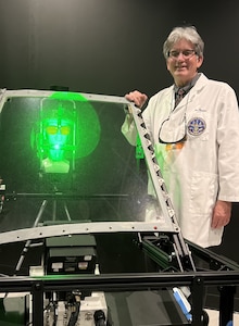 Man in lab coat stands in a research lab next to a laser veiling glare simulator while a green laser is projected onto the veil.