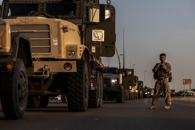 A Marine in battle equipment stands next to a military vehicle at twilight.