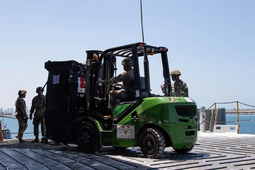 Soldiers operate a forklift carrying a pallet of humanitarian assistance cargo on a pier.