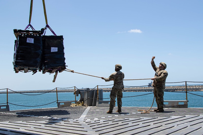 Two soldiers use a rope to guide a pallet containing humanitarian assistance cargo onto a ship. The pallet is suspended by straps.
